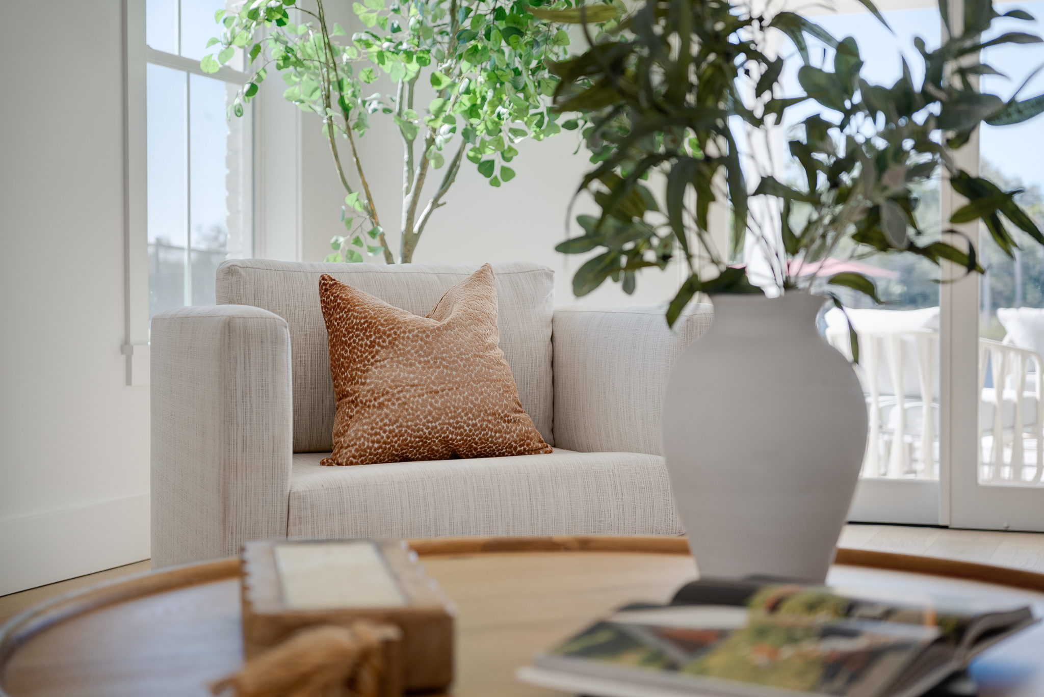 A beige armchair with a textured brown cushion sits near a window and large leafy plant; a round wooden coffee table with a vase and open book is in the foreground.