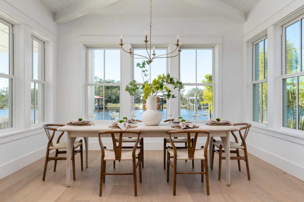 Bright dining room with a wooden table set for six, modern chairs, a white vase with greenery, large windows, and a view of trees and water outside.