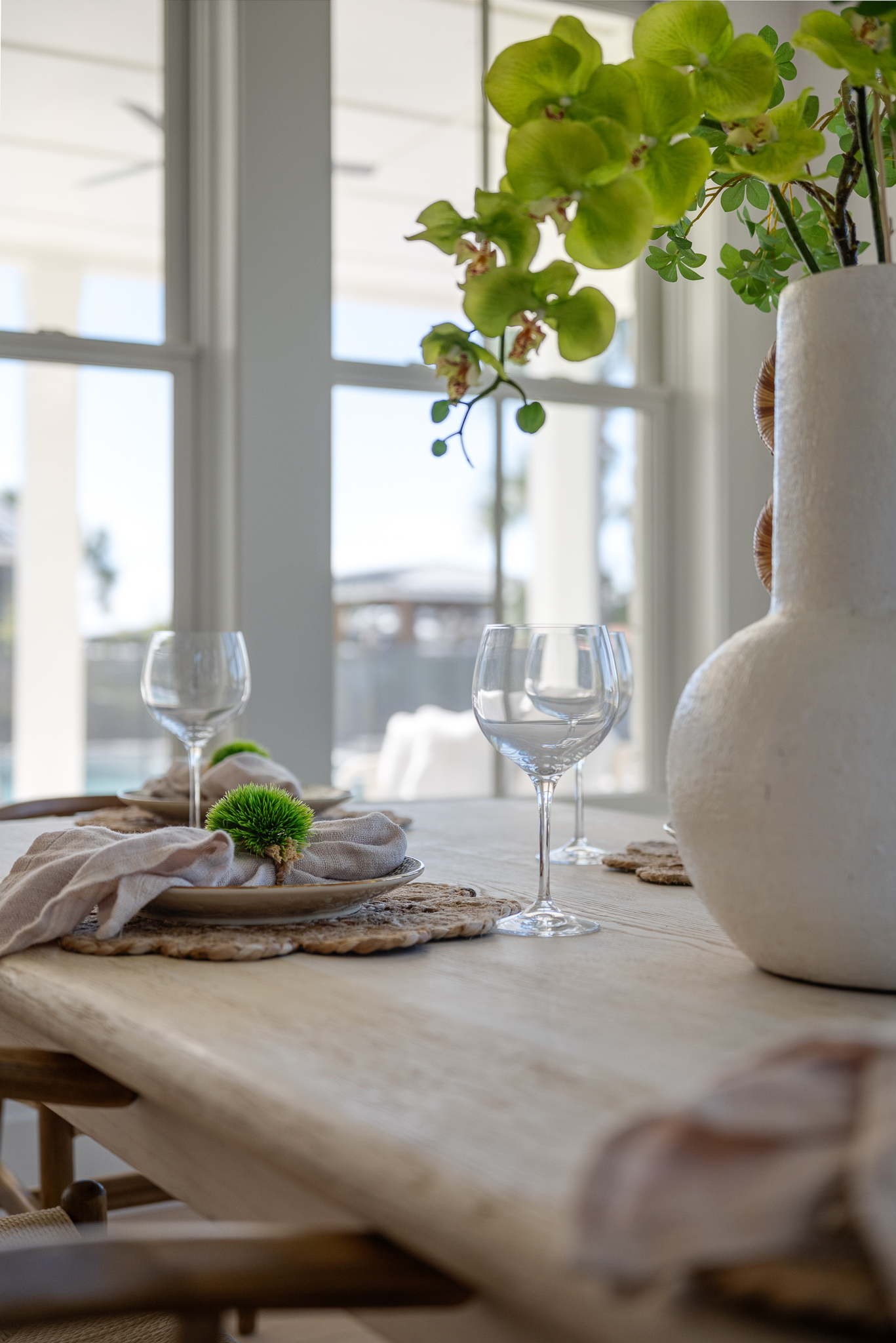 A wooden dining table set with woven placemats, napkins, empty wine glasses, and a white vase with green flowers, positioned near large windows.