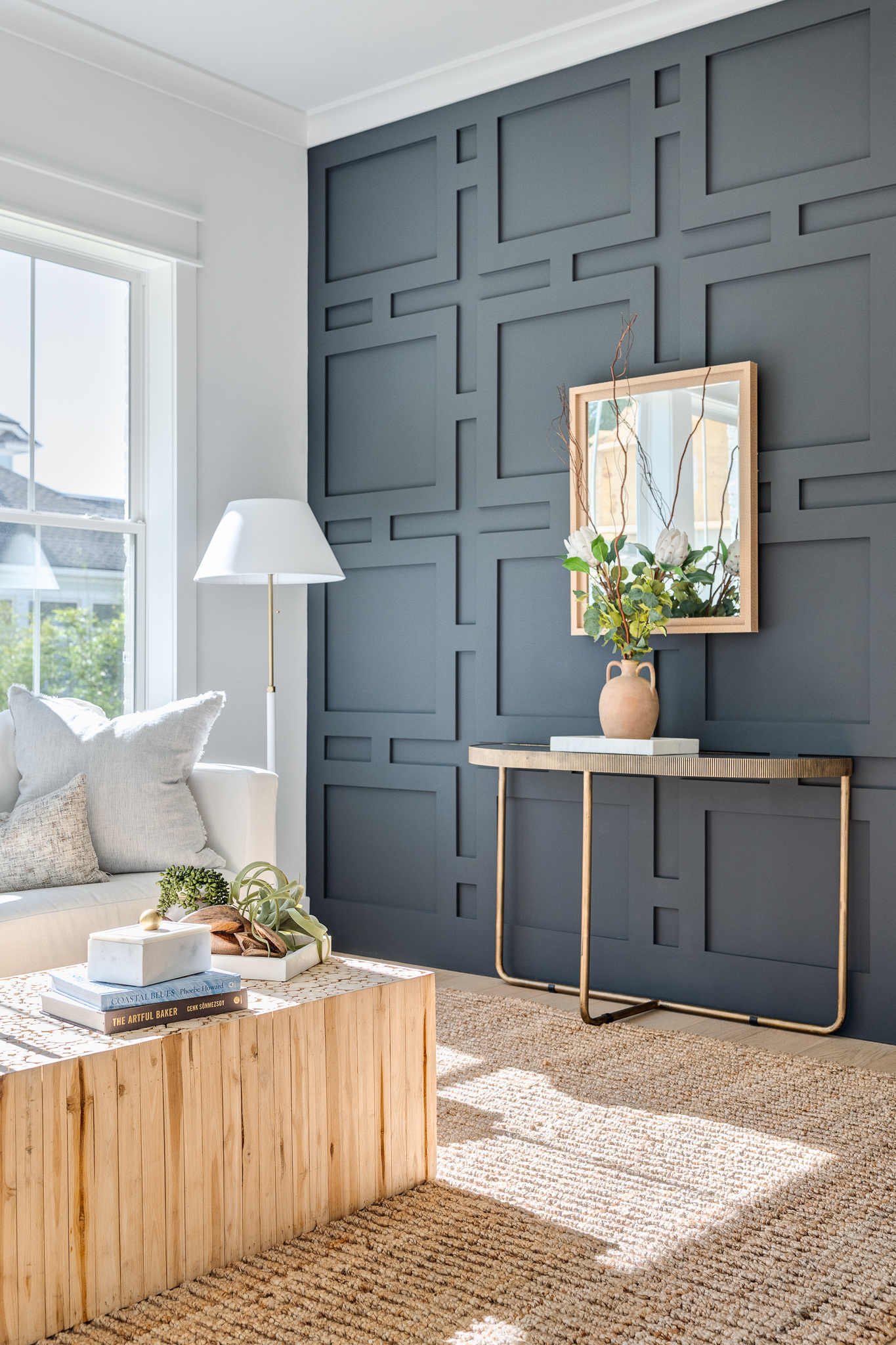 Modern living room with a geometric dark gray accent wall, a console table with a vase and mirror, a white sofa, and a wooden coffee table with books. Natural light streams in.