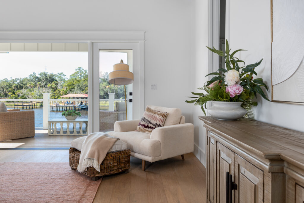 A cozy armchair with a throw blanket sits by a large window and door leading to a waterfront patio; a cabinet with a floral arrangement is on the right.