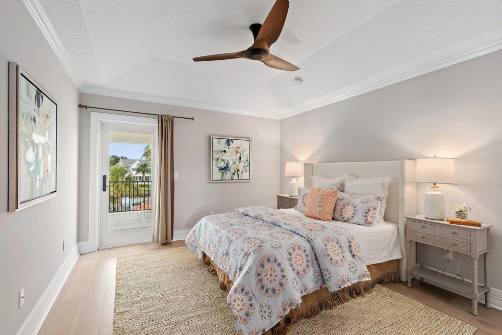 A modern bedroom with a large bed, two nightstands with lamps, abstract art on the walls, a ceiling fan, and a door leading to a balcony with a view of palm trees.